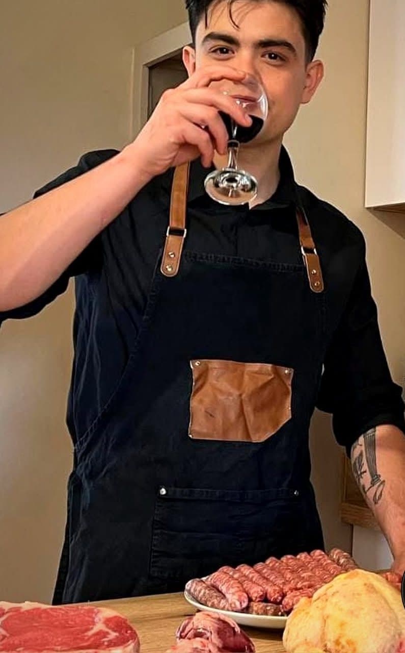 Peter, le gérant de Carnis, en tablier noir à bretelles de cuir, un verre de vin rouge à la main, devant une côte de bœuf, des saucisses et des volailles